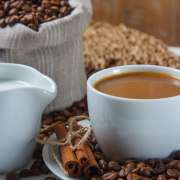 close-up-cup-coffee-with-coffee-beans-sack-saucer-milk-dry-cinnamon-trivet-white-surface-vertical